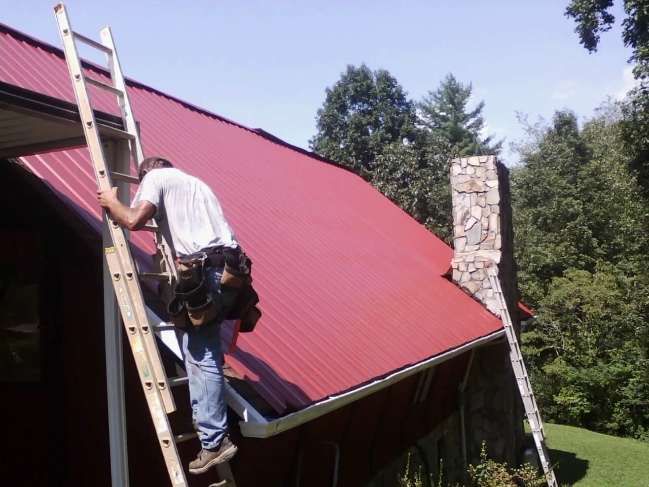 Roofer on a ladder beside a red metal roof and stone chimney — Soffit & Fascia Repair by Roofing Brothers in Delcambre